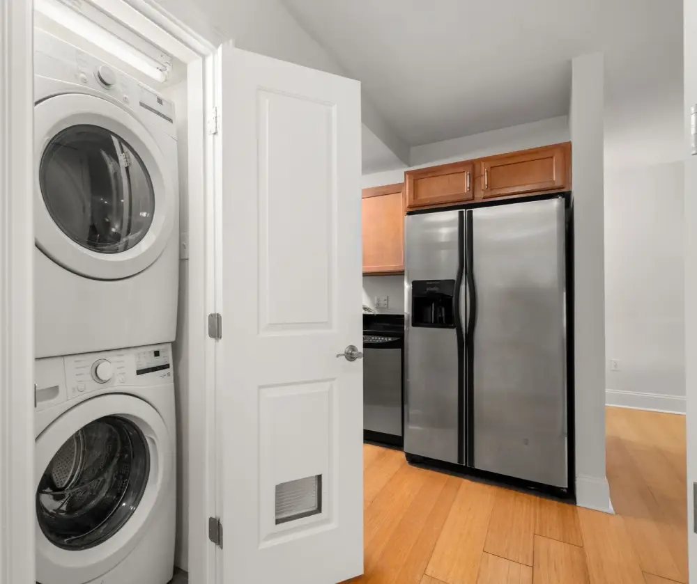 Stacked white washer and dryer inside a small laundry closet with a door, next to a kitchen area with hardwood floors and stainless steel refrigerator.