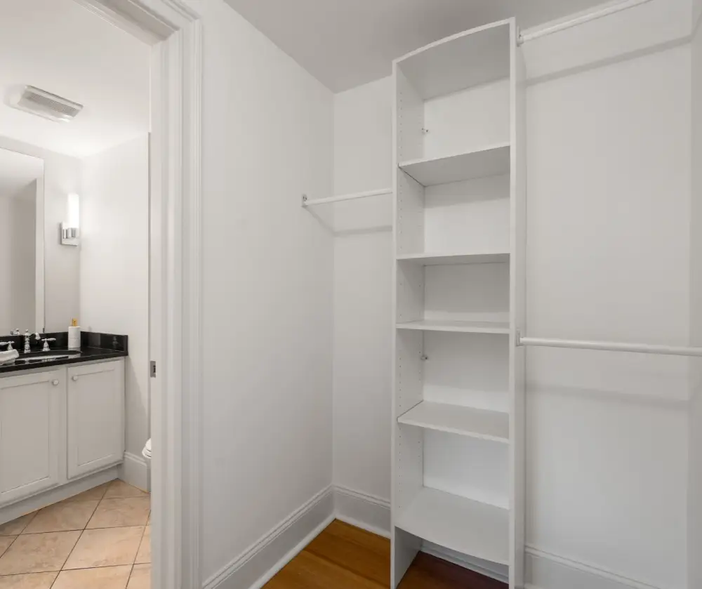 Empty white walk-in closet with built-in shelves and hanging rods, adjacent to a bathroom with a black countertop.