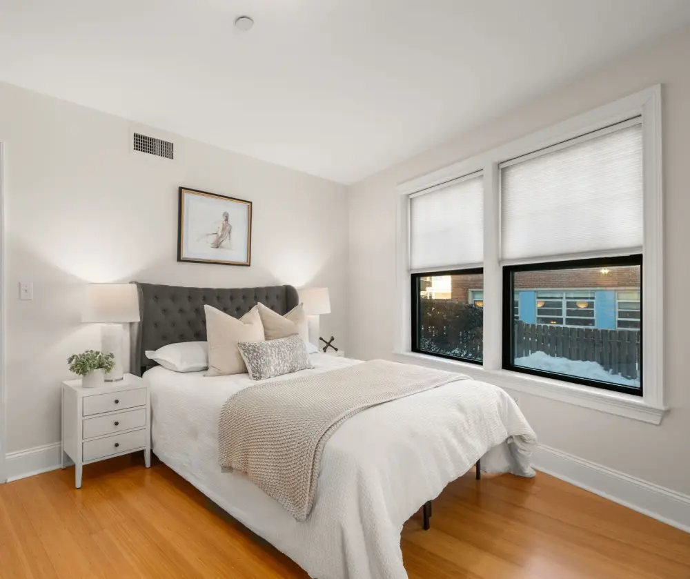 Cozy bedroom with white walls, a bed with a gray tufted headboard, beige and patterned pillows, a knitted throw, two bedside tables with lamps, and large windows with white blinds.