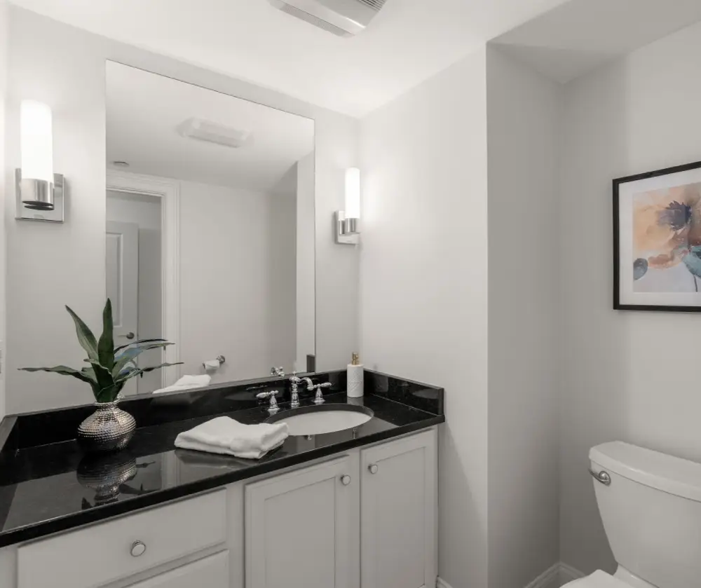 Modern bathroom with a black granite countertop, white cabinets, an oval sink, a plant in a silver vase, a towel, wall-mounted sconces, a toilet, and a framed abstract artwork.