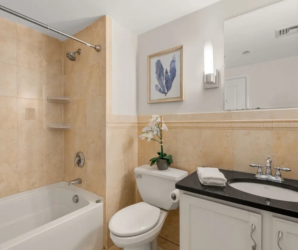 Bathroom with a white bathtub, tiled shower area, white toilet with a potted orchid on top, and a black countertop sink under a large mirror.