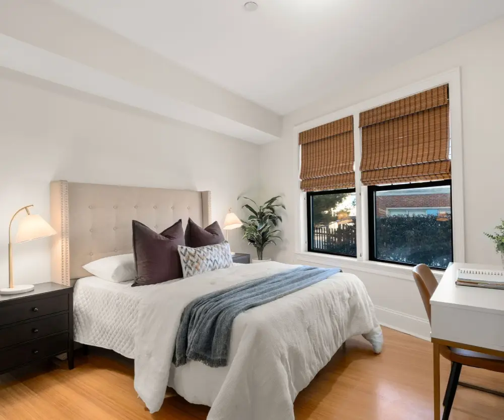 Bright bedroom with a beige tufted headboard bed, white bedding, dark pillows, a blue throw blanket, wooden blinds on two windows, and a white desk with a brown chair.