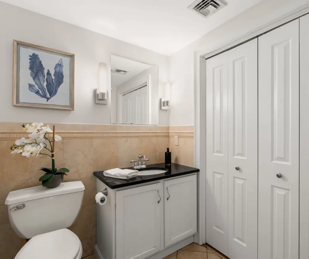 Modern bathroom with white toilet, white vanity with black countertop, wall mirror, and closet with double doors.