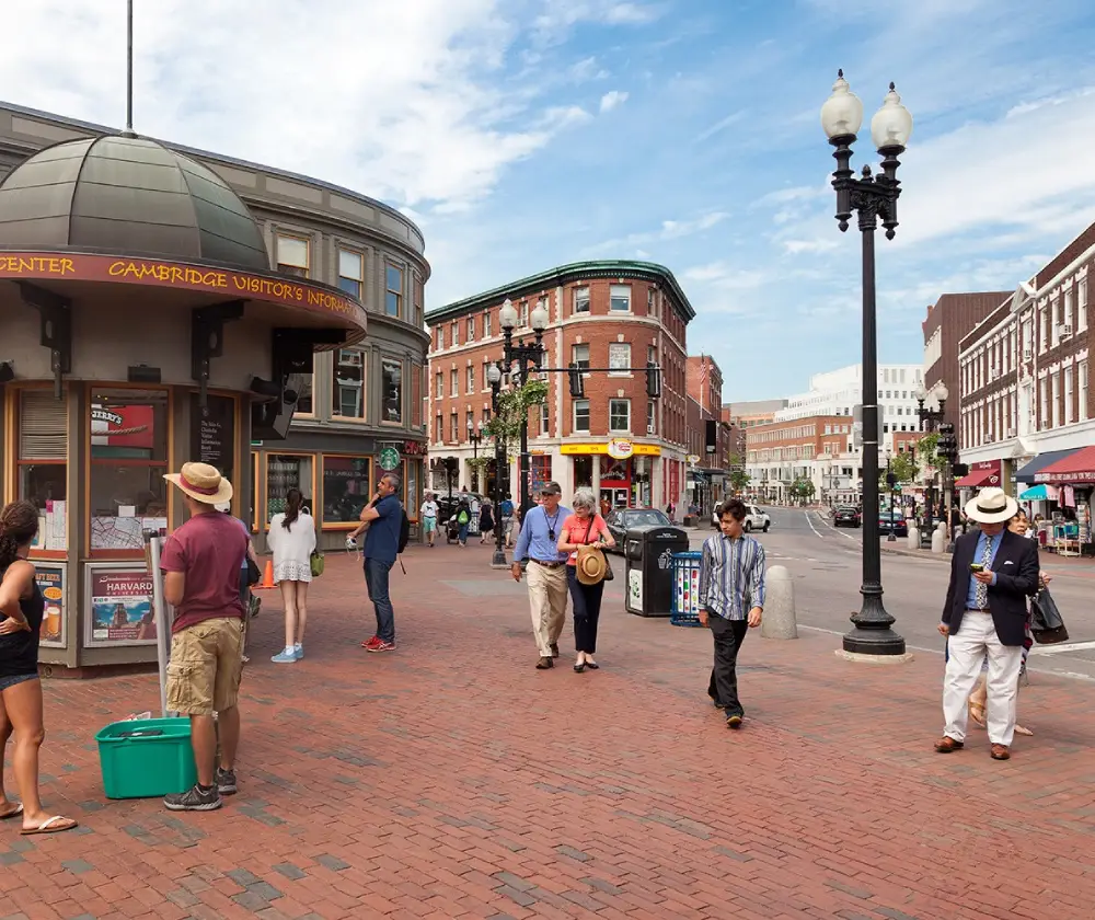 Street scene at Harvard Square in Cambridge, Massachusetts, with pedestrians walking on brick pavement near shops and a visitor information center.