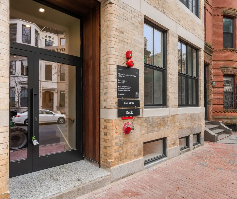 Entrance with glass doors numbered 144 next to a brick building with signs for parking and deck and contact information for real estate agents.