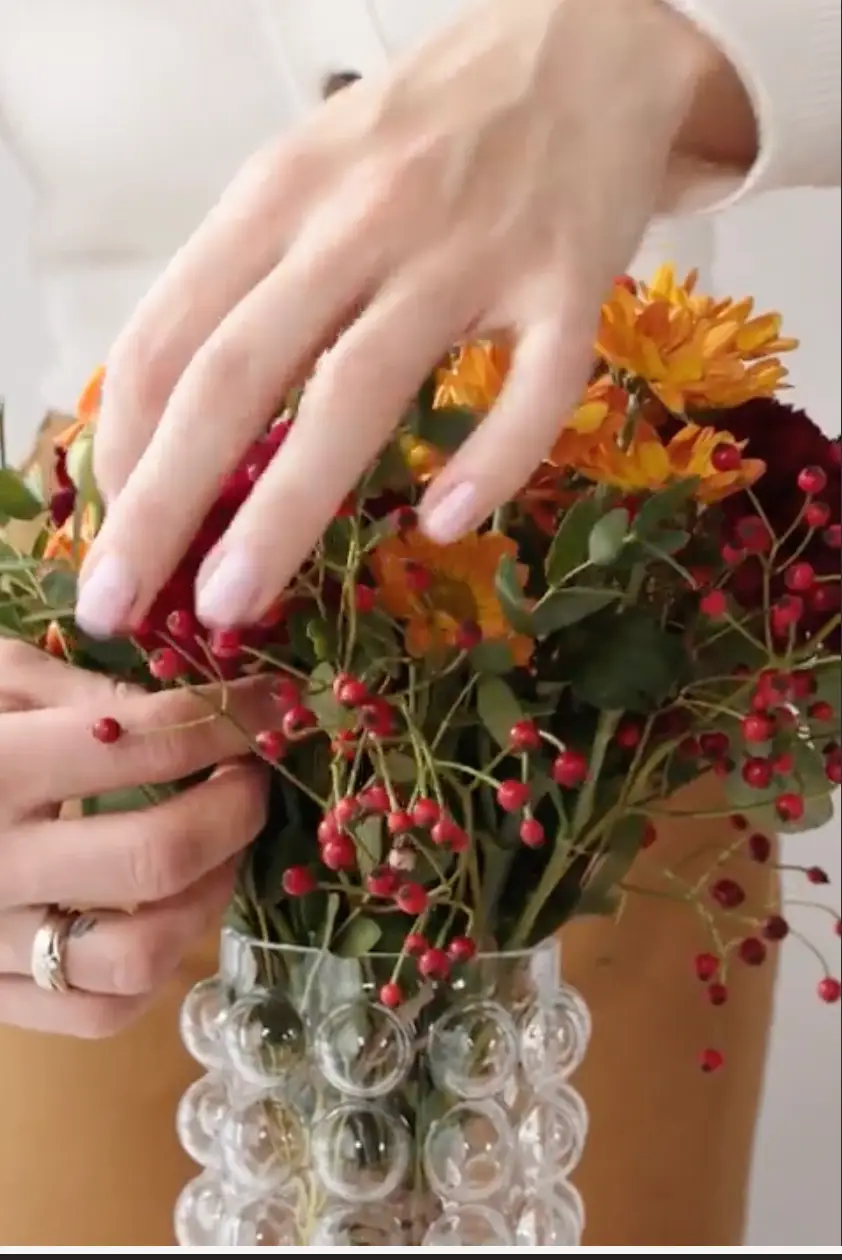 Hands arranging a bouquet of orange and yellow flowers with red berries in a clear textured vase.