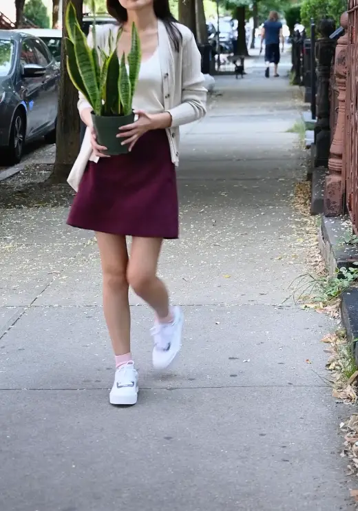 Person walking on a sidewalk holding a potted snake plant, wearing a beige cardigan, white top, burgundy skirt, and white sneakers.