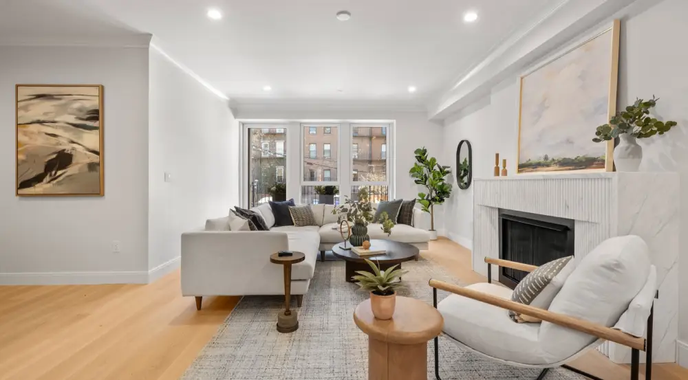 Bright modern living room with white sectional sofa, round coffee table, armchair, fireplace, and large window overlooking buildings.