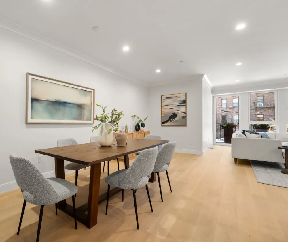 Modern dining area with a wooden table, five gray upholstered chairs, abstract wall paintings, and a bright living space with large windows.