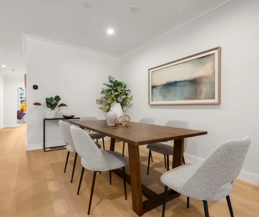 Modern dining room with six gray upholstered chairs around a dark wooden table adorned with two white vases and a decorative orb, light wood floor, and abstract wall art.
