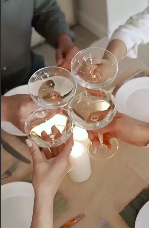 Four people clinking clear cocktail glasses filled with a light beverage over a dining table set with white plates and cutlery.