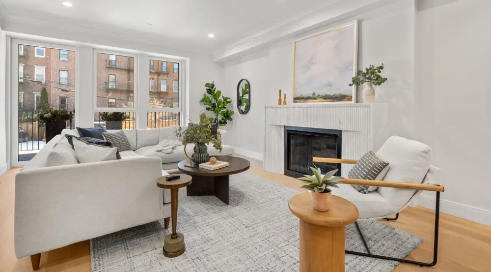 Bright living room with a white sectional sofa, a white armchair with wooden armrests, a gray rug, round wooden coffee tables with plants, and a fireplace topped with a landscape painting.