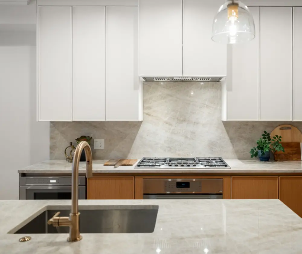 Modern kitchen with marble countertops, stainless steel sink with brass faucet, gas stove, white upper cabinets, and wooden lower cabinets.
