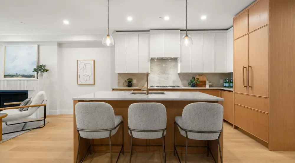 Modern kitchen with a marble island and three grey upholstered chairs, white cabinets, wooden built-ins, and pendant lights.