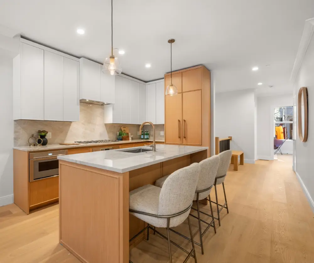 Modern kitchen with wooden cabinets, white upper cabinets, marble countertop island with two upholstered bar stools, and pendant lights.