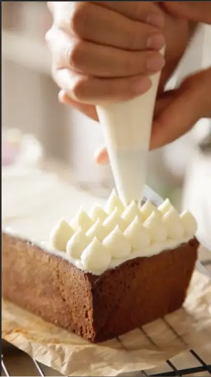 Close-up of hands decorating a rectangular chocolate cake with white frosting using a piping bag.