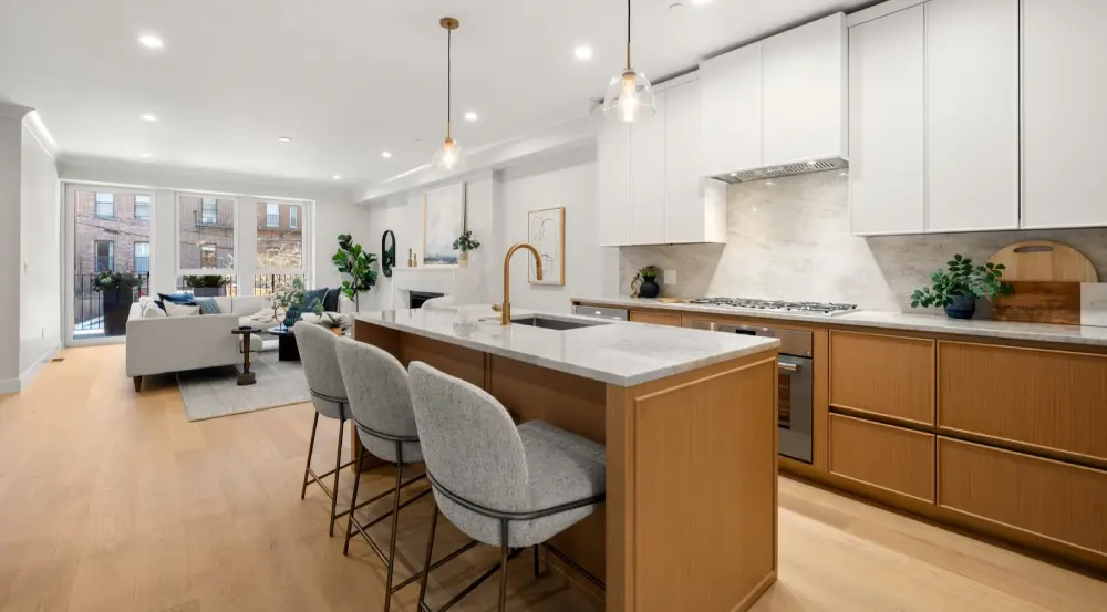 Modern open-concept kitchen with wooden cabinetry, white countertops, three upholstered bar stools at the island, and a living area with a sofa and large windows in the background.