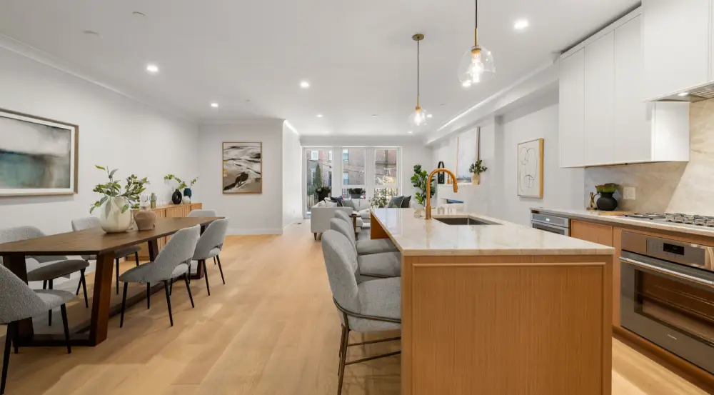 Open-concept modern living space with a wood dining table and chairs on the left, a kitchen island with gray chairs in the center, and a seating area with sofas near large windows in the background.