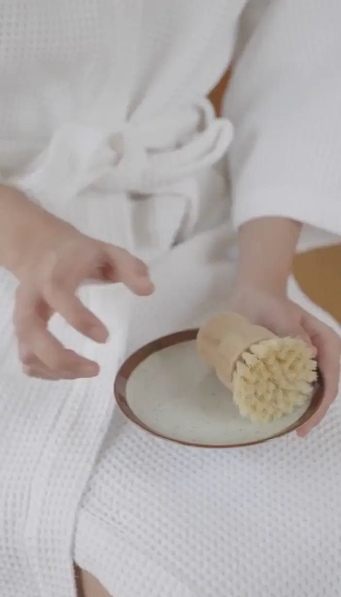 Person in white waffle-textured robe holding a round brush on a small plate.