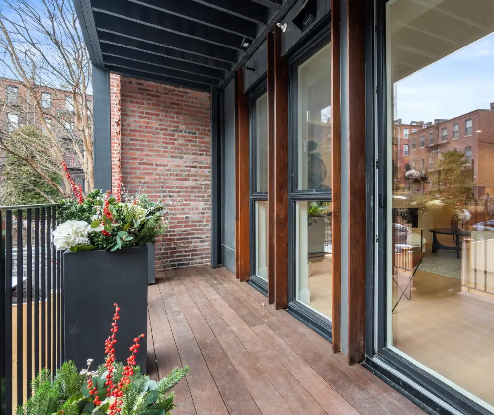 Wooden balcony with black railing, potted plants with red and white flowers, and glass doors with wooden frames leading inside.