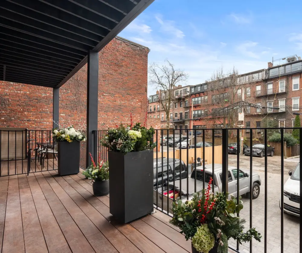 Covered balcony with wooden floor, black metal railing, potted floral arrangements, overlooking a parking lot and urban buildings.