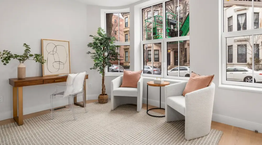 Bright sitting area with two white armchairs featuring rose-colored pillows, a small round wooden table, a tall potted plant, and a wooden desk with a clear chair and decorative items.