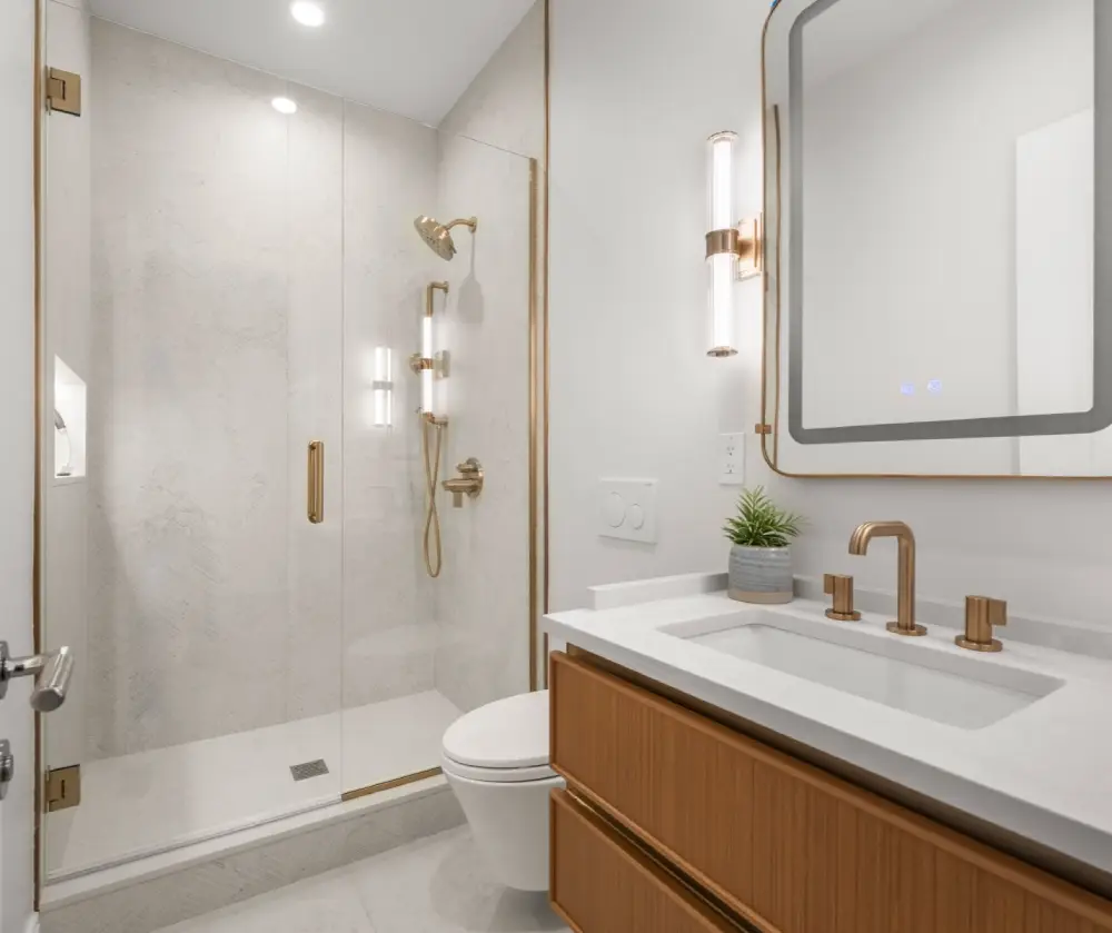 Modern bathroom with a glass shower enclosure, gold fixtures, wooden vanity with white countertop, rectangular mirror, and small potted plant.
