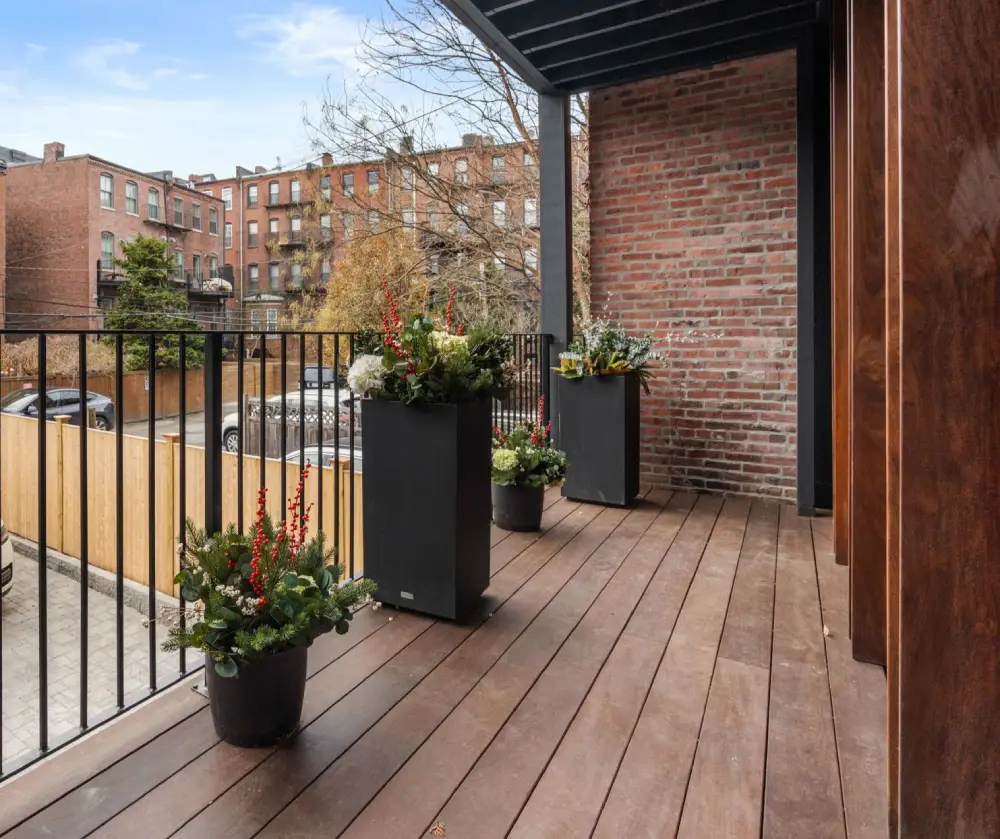 Wooden balcony with black metal railing, four planters containing green and red plants, and a brick wall background under a roof.