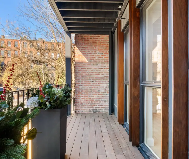 A bright balcony with wooden flooring, dark metal railings, potted greenery with red berries, and large windows with wood frames, against a brick wall and clear sky.