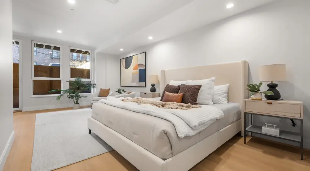 Modern bedroom with a beige upholstered bed, white bedding, decorative pillows, two bedside tables with lamps and plants, a large white area rug, and large windows letting in natural light.