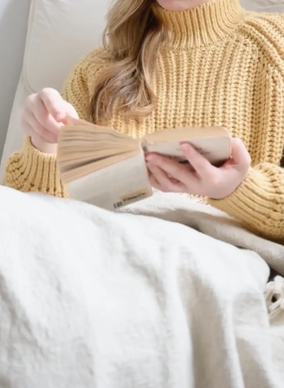 Person in a yellow knitted sweater reading a book while covered with a light blanket.