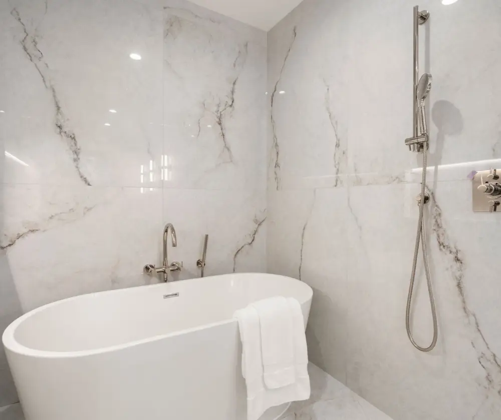 Modern bathroom with a white freestanding bathtub, marble-patterned walls, and a chrome showerhead with a handheld attachment.