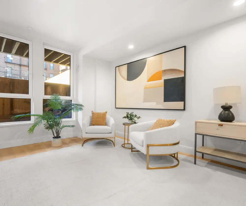 Modern living room with two white armchairs with gold frames and orange pillows, large abstract wall art, potted plant, and a side table with a lamp.