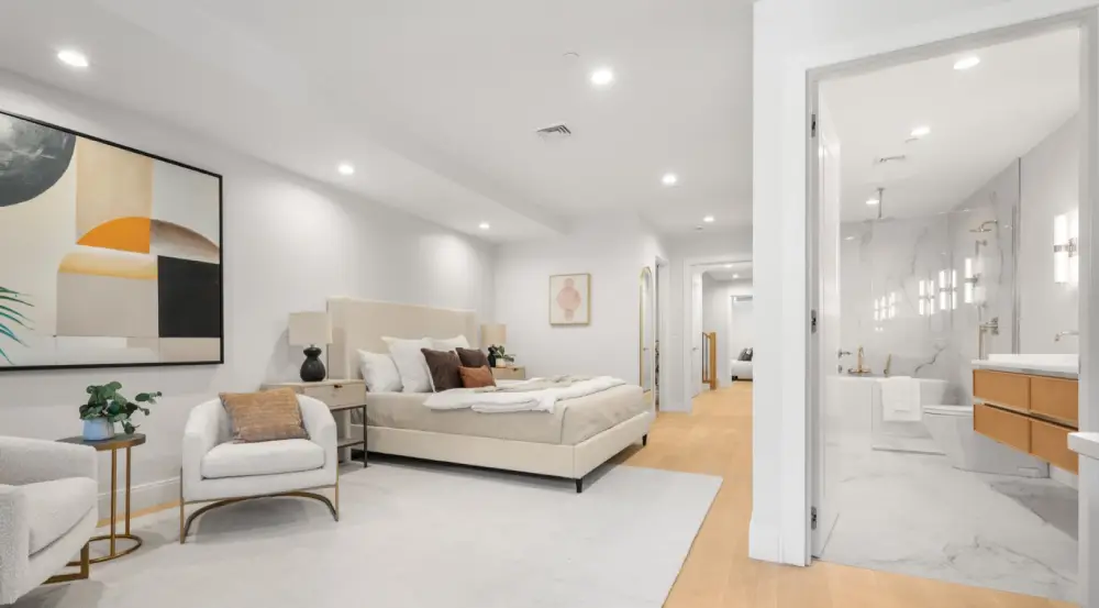 Modern bedroom with beige upholstered bed, white armchairs, abstract wall art, and an open door to a bright bathroom with marble walls.