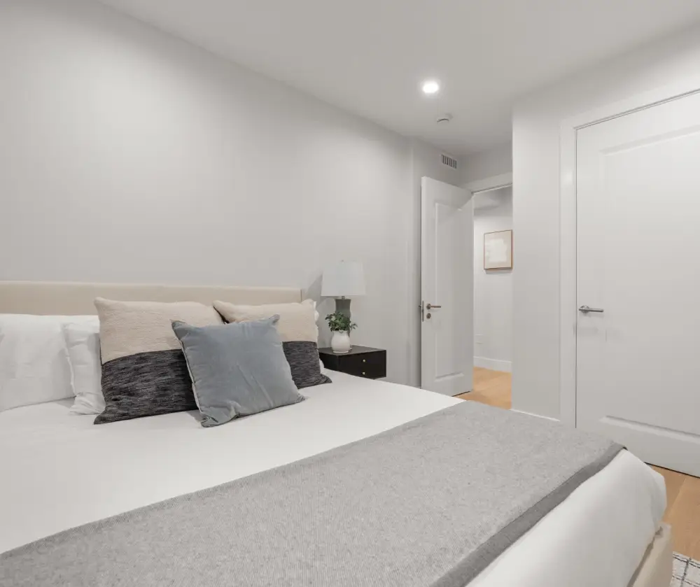 Minimalist bedroom with a bed featuring white and gray pillows, a gray throw blanket, a bedside table with a lamp and plant, and two doors on white walls.