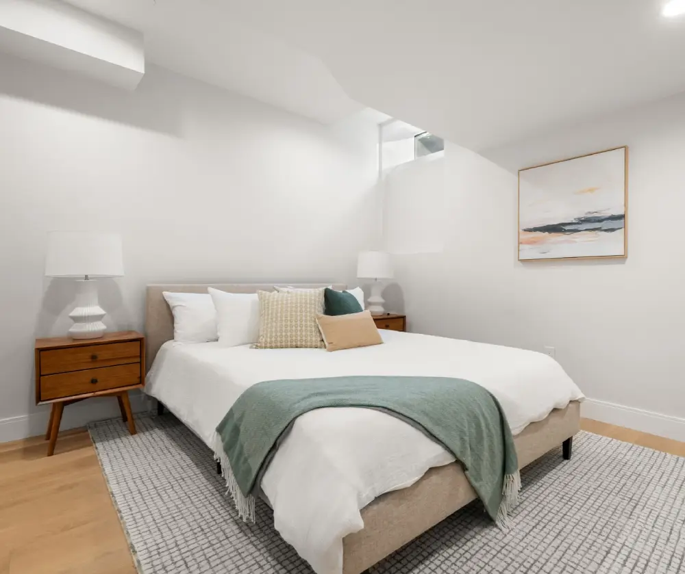 Minimalist bedroom with a beige upholstered bed, white bedding, green throw, two wooden nightstands with white lamps, and an abstract painting on the wall.