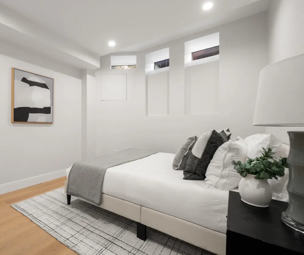 Minimalist bedroom with a bed featuring white and gray pillows, a gray throw blanket, a black nightstand with a white lamp and small plant, light wood flooring, and abstract wall art.