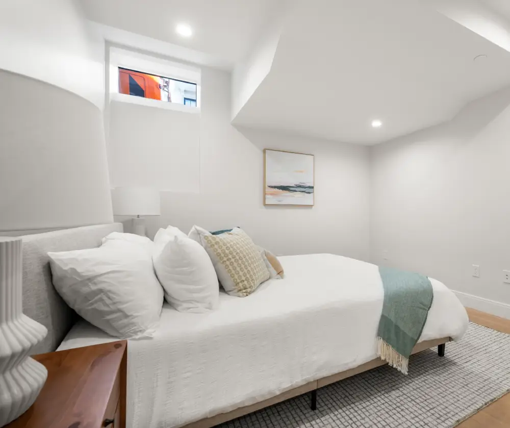 Minimalist bedroom with white walls, a bed with white bedding and decorative pillows, a green throw blanket, a wooden nightstand with a white lamp, a framed abstract painting, and a small window near the ceiling.
