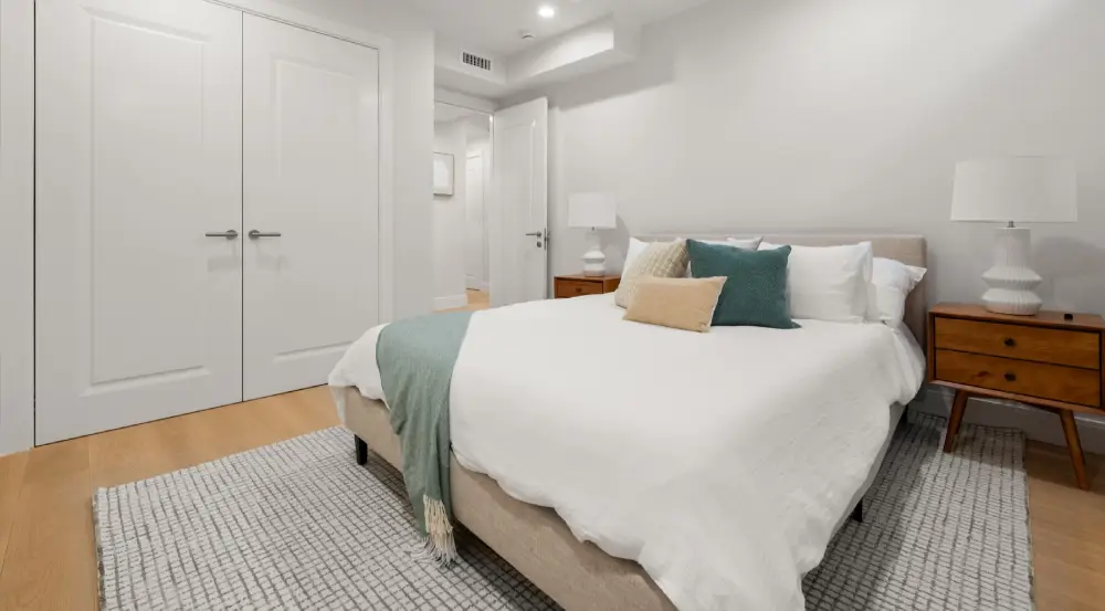 Minimalist bedroom with beige bed, white linens, decorative pillows, green throw, wooden nightstands with white lamps, white walls, and closed double doors.