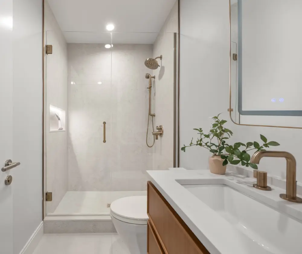 Modern bathroom with glass-enclosed shower, gold fixtures, white countertop, wooden vanity, and small potted plant.