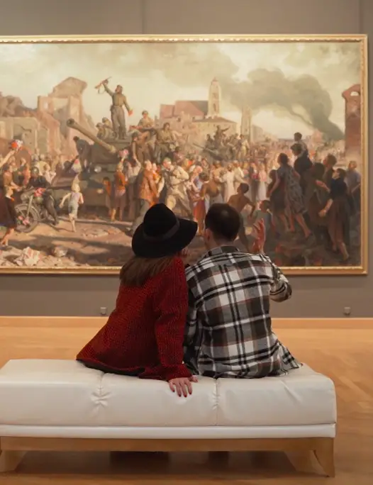 Couple sitting on a white bench in an art gallery, viewing a large historical painting depicting a crowd around a tank amid ruins.