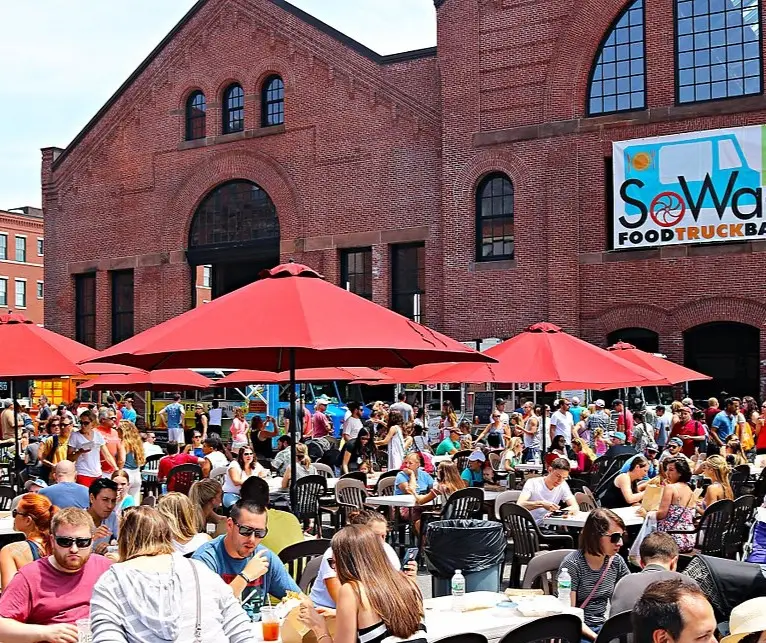 Crowded outdoor food truck event with people sitting under red umbrellas and a brick building in the background.