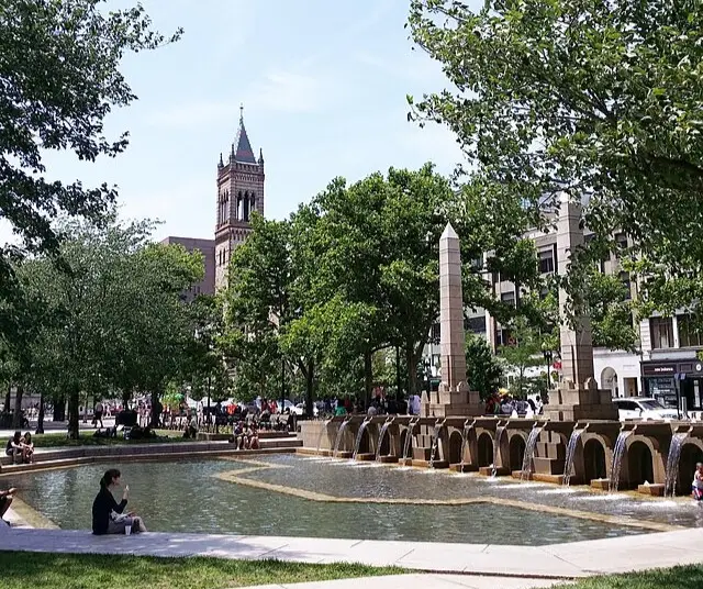 Fountain with cascading water and an obelisk in a park surrounded by trees and buildings, with people sitting and walking nearby.