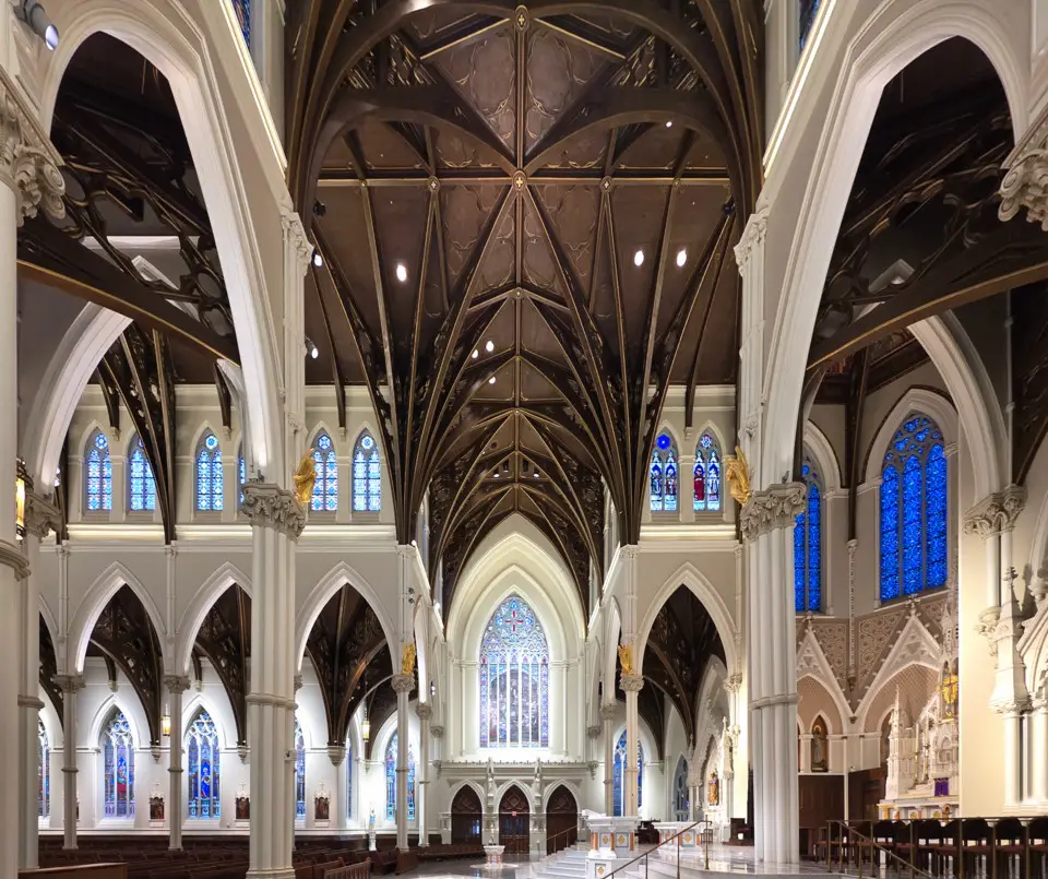 Interior view of a cathedral transept showcasing Gothic-style pointed arches, dark wooden vaulted ceiling beams, and stained glass windows.
