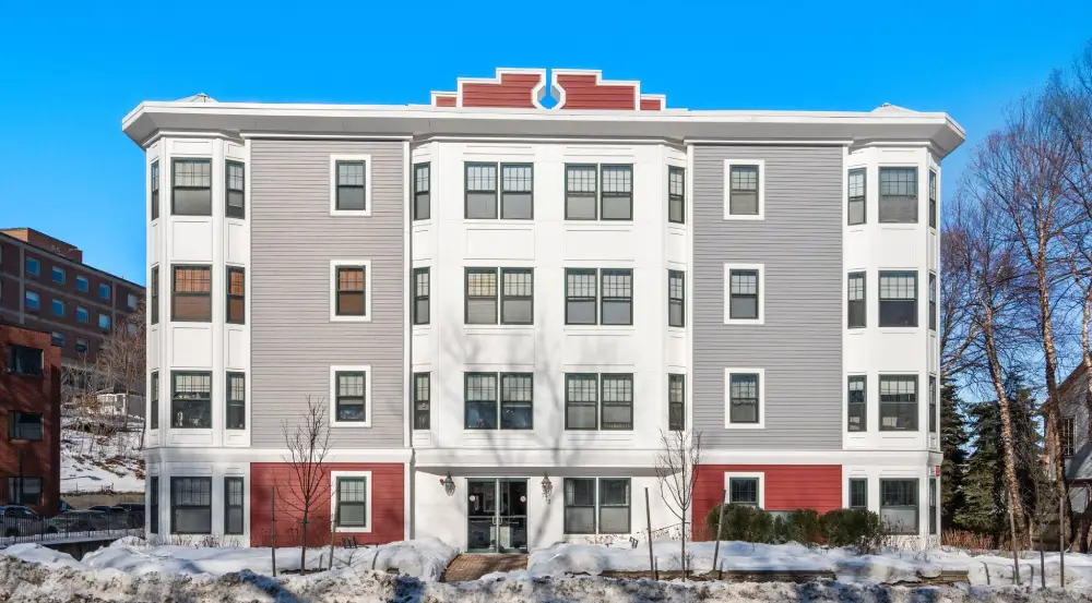 Four-story apartment building with gray, white, and red facade surrounded by snow under a clear blue sky.