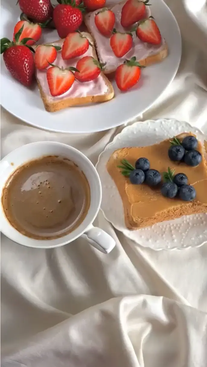 Cup of coffee, toast with peanut butter and blueberries, and toast with strawberry yogurt and sliced strawberries on white plates over white fabric.