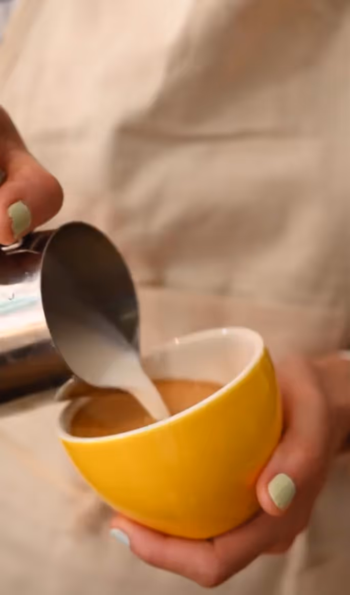 Person pouring steamed milk from a metal pitcher into a yellow cup of coffee.