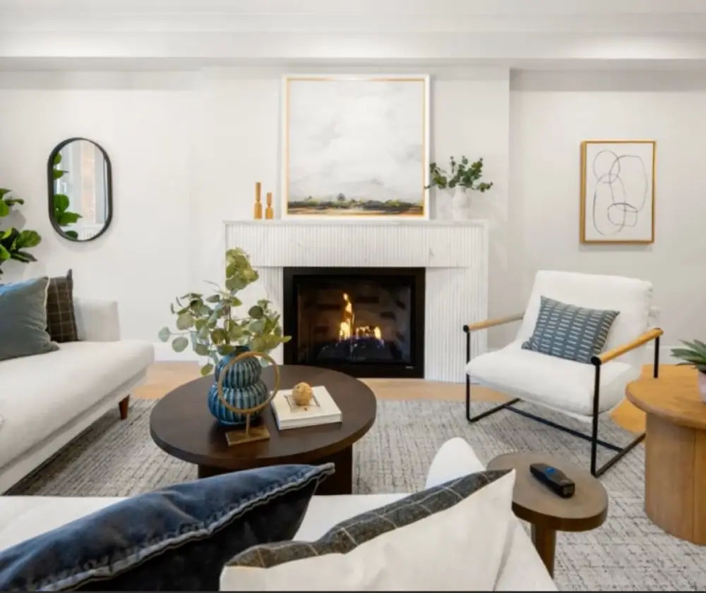 Modern living room with white walls, a fireplace, a round dark wood coffee table with blue vase and books, white armchair with blue cushion, and decorative wall art.