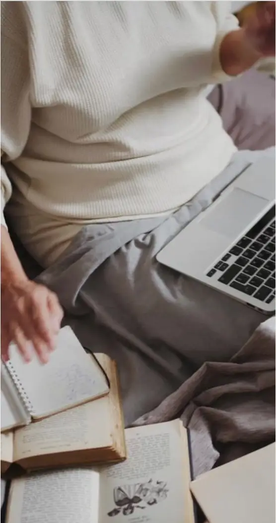 Person wearing a white sweater sitting on a bed with a gray blanket, using a laptop and writing in a notebook surrounded by open books.