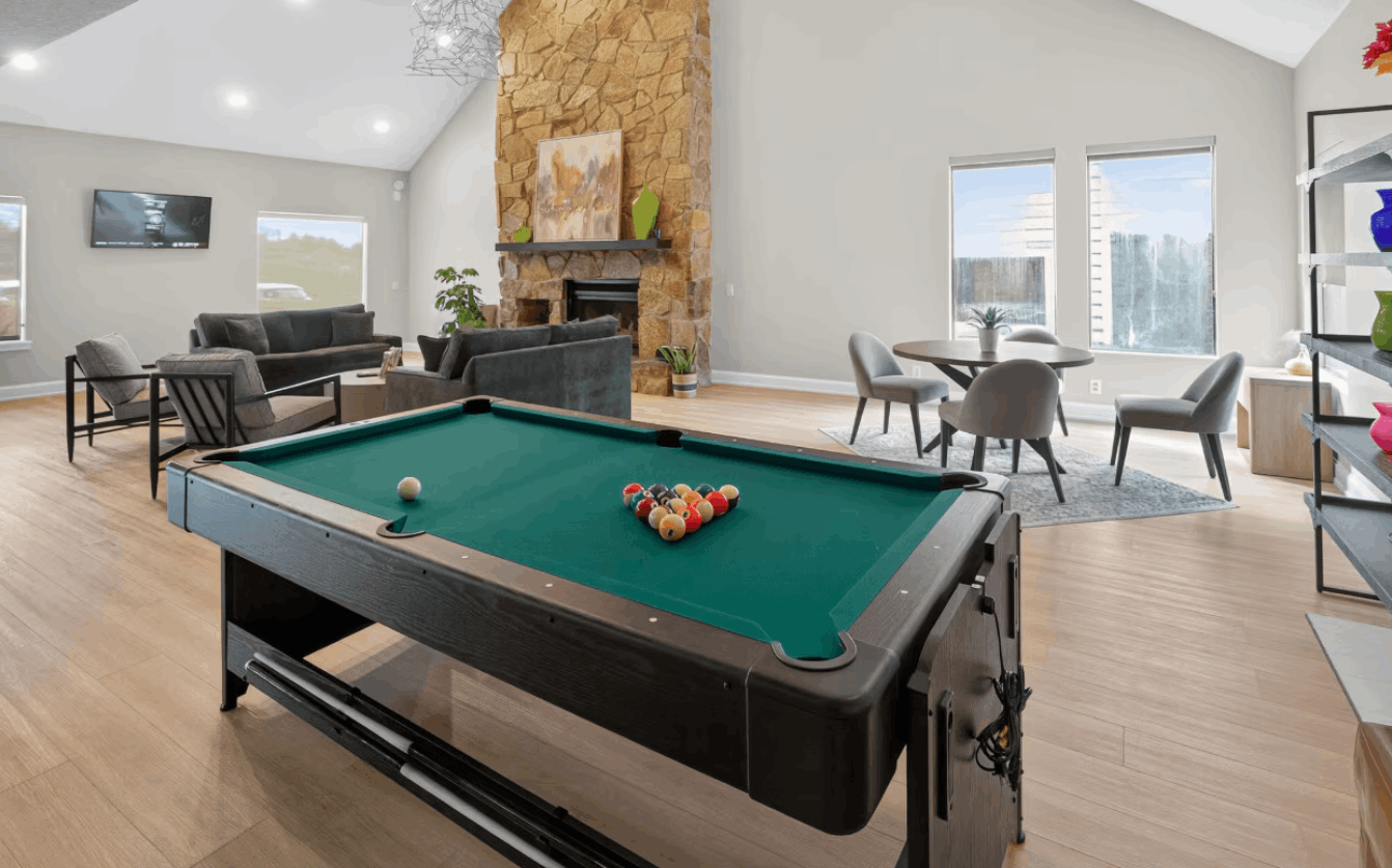 Modern game room with a green pool table, gray sofas, a stone fireplace, and a round table with four chairs near windows.
